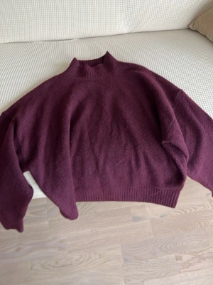 Vinröd oversized stickad tröja - Mysig vinröd stickad tröja med hög hals och avslappnad passform. Tröjan har ribbade muddar vid hals, ärmslut och nederkant. Perfekt för lager-på-lager och en chill look.