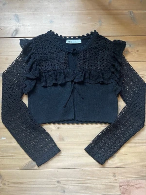Svart spets kofta/topp från Zara - Svart blus från Zara med långa ärmar i spets och volangdetaljer över axlar och bröst. Blusen har knytning framtill och är tillverkad i ett genombrutet spetsmaterial som ger en cool och feminin look.