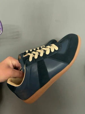 Masion margiella skor - Snygga sneakers i mörkblått läder och mocka med beige snörning och klassisk gummisula. Skorna har en stilren design med lågt skaft och passar perfekt till streetwear eller casual outfits.