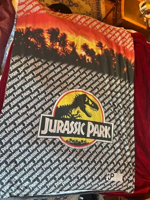 Jurassic Park påslakan retro - Säljer ett retro påslakan med Jurassic Park-tryck. Motivet har solnedgång, palmer och det klassiska Jurassic Park-loggan i gult, rött och svart. Materialet är mjukt tyg, troligen bomull. Perfekt för dig som älskar filmklassiker och coola prints.