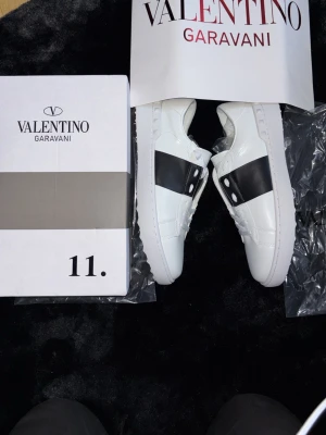 Valentino Garavani vita sneakers - Snygga vita sneakers från Valentino Garavani med en bred svart rem över mitten och coola nitar på sidorna. Skorna är i läder och har en klassisk låg profil med vita snören och diskret branding. Perfekta för dig som vill sticka ut med stil.