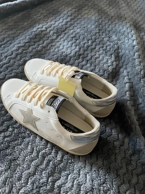 Golden Goose vita sneakers med silverdetaljer - Golden Goose Super-Star sneakers i vitt läder med silverfärgad häl och ikonisk stjärna på sidan. Skorna har vita snören, svart logotyp på plösen och en klassisk låg siluett. Perfekta för dig som gillar stilrena och trendiga sneakers. Dem är helt nya bara provade. Har dustbag men vet ej vart den ligger, kan pröva att hitta den. Säljer för skorna var inte min storlek.