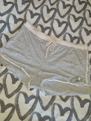 Shorts - Snygga grå shorts med vit snörning i midjan och vit kant längs benen. På ena benet finns en broderad stjärna med regnbågssvans. Perfekta för sommaren och chill dagar. Köpte för väldigt längesen så väl använda men inga synliga defekter.