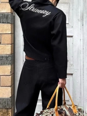 Svart jeansjacka från StreetWearEazy  - Snygg svart jeansjacka från StreetWearEazy med dragkedja framtill och vit brodyrtext 'StreetWearEazy' på ryggen. Jackan har en minimalistisk look med diskret logga vid nederkanten och raka ärmar. Perfekt för dig som gillar streetwear och vill ha en clean stil.