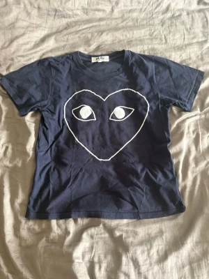 Mörkblå Comme des Garçons PLAY t-shirt - Mörkblå t-shirt från Comme des Garçons PLAY med det ikoniska hjärtmotivet och ögon i vitt på bröstet. Klassisk passform med rund hals och korta ärmar. Tillverkad i mjuk bomull som känns skön mot huden.