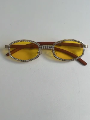 Gula solglasögon med strasskant - Unika solglasögon med rektangulära, gula glas och glittrande strass runt hela bågen. Skalmarna är bruna och bågen har en cool vintagekänsla. Perfekt accessoar för dig som vill sticka ut och addera lite extra bling till din stil.
