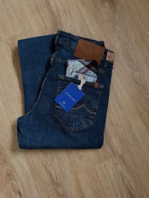 Mörkblå jeans från Jacob Cohen - Snygga mörkblå jeans från Jacob Cohen med klassisk femficksmodell och raka ben. Jeansen har detaljer som bruna läderpatches, silverfärgade knappar och kontrastsömmar. Tillverkade i mjuk denim med exklusiv känsla och diskret logotyp på bakfickan.