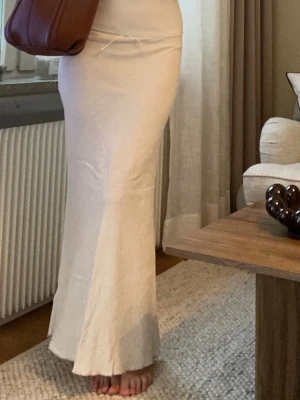 Beige långkjol  - Superfin långkjol i en mjuk beige/cremefärg från Urban outfitters. Den har en figurnära passform som faller snyggt längs kroppen och passar perfekt både till vardag och lite mer uppklädda tillfällen.