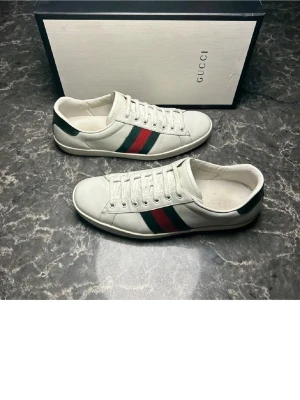 Vita Gucci sneakers med ränder - Säljer ett par klassiska vita sneakers från Gucci med de ikoniska gröna och röda sidoremsorna. Skorna har låga skaft, vita snören och är tillverkade i slätt läder. ej box.Perfekta för dig som vill ha en clean och lyxig look med streetvibe.