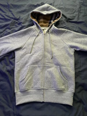 Grå zip hoodie från Burberry - Snygg grå zip hoodie från Burberry med klassiskt rutigt foder i huvan. Hoodien har dragsko i huvan, två stora fickor fram och ribbade muddar. Tillverkad i mjuk bomullsblandning för extra komfort. Perfekt för en avslappnad och trendig look.
