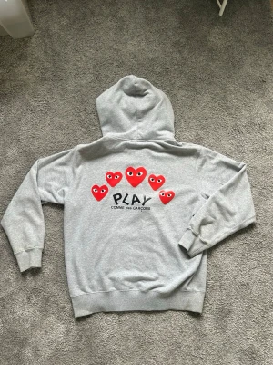 Grå hoodie Comme des Garçons Play - Snygg grå zip-hoodie från Comme des Garçons Play med röda hjärtan och logga på ryggen samt ett litet rött hjärta på bröstet. Klassisk huva, fickor fram och ribbade muddar. Perfekt för en chill och trendig look.