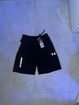 Svarta shorts från Under Armour - Snygga svarta shorts från Under Armour med färgglad logga på benet och vita detaljer. Shortsen har elastisk midja med snörning och är perfekta för träning eller chill. Tillverkade i mjukt och lätt syntetmaterial för maximal komfort.