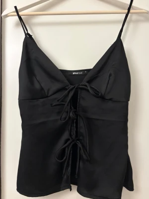 Svart snörad linnetopp Gina Tricot - Snygg svart linnetopp från Gina Tricot med tunna axelband och snörning framtill. Toppen har en v-ringad skärning och är tillverkad i ett glansigt, satinliknande material som ger en lyxig känsla. Perfekt för att lyfta vilken outfit som helst.