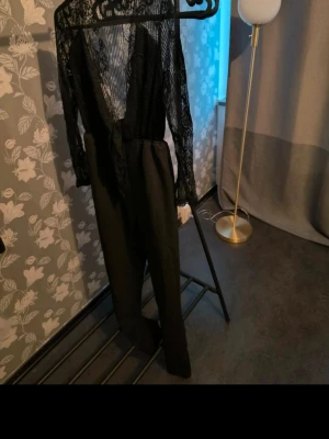 Svart spetsjumpsuit - Snygg svart jumpsuit med långärmade spetsdetaljer och djup v-ringning. Byxdelen är i ett slätt tyg med normal passform. Perfekt för dig som vill sticka ut.