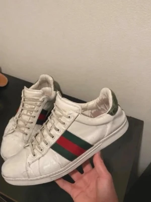 vita sneakers med gröna detaljer - Snygga vita sneakers  i läder med klassiska gröna och röda ränder på sidan. Baksidan har en grön detalj med krokodilstruktur. Skorna har vita snören och en robust sula med mönster. Perfekta för dig som vill ha en ikonisk och trendig look hör av dig vid minsta lilla fundering .