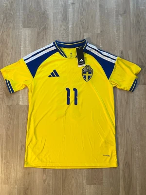 Sverige Elanga VM-2026 Fotbollströja - Gul och blå Sveriges Hemma fotbollströja med Elanga och nummer 11 på ryggen. Tröjan har korta ärmar och broderad landslagssköld. Tillverkad i lätt och ventilerande material, perfekt för fotbollsträning eller match. HELT NY