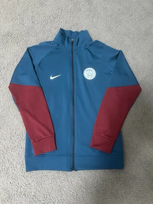 PSG träningsjacka blå vinröda ärmar - Snygg PSG träningsjacka från Nike i blått med vinröda ärmar och hel dragkedja. Jackan har PSG-logga på bröstet och vit Nike Swoosh. Perfekt för fotbollsträning eller som sportig streetwear. Har hela setet om någon vill köpa det. Priset kan diskuteras! Byxorna blir 300kr till! 