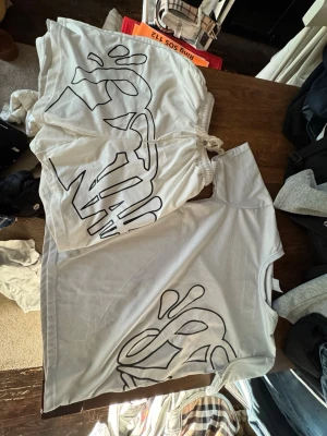 Vit t-shirt och shorts med grafiskt tryck - Säljer en vit t-shirt med ett stort svart grafiskt tryck framtill. T-shirten har korta ärmar och en avslappnad passform. Perfekt för dig som gillar streetwear och vill sticka ut med en unik design.