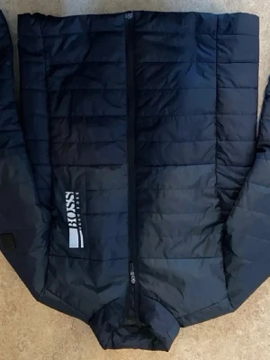 Svart pufferjacka från Boss - Säljer en svart pufferjacka från Boss med quiltad design och logotyp på bröstet. Jackan har hög krage och dragkedja framtill. Perfekt för kalla dagar och har en modern, sportig look.