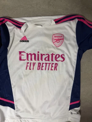 Arsenal träningsset Adidas blå/vit - Säljer ett Arsenal träningsset från Adidas med matchtröja i vitt med rosa och marinblå detaljer samt klubbmärke och sponsortryck. Tillhörande shorts i marinblått med rosa ränder och Arsenal-logga. Materialet är lätt och andas, perfekt för fotbollsträning.