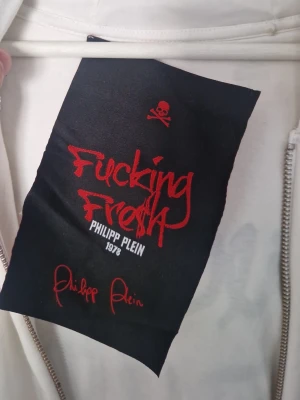 Vit hoodie Philipp Plein med tryck - Säljer en vit hoodie från Philipp Plein med stort svart tryck 'Fucking Fresh' på ryggen och coola slitningar över hela plagget. Hoodien har huva, dragkedja och ribbade muddar. Märkesdetaljer och edgy streetstyle-vibe. Priset kan även diskuteras