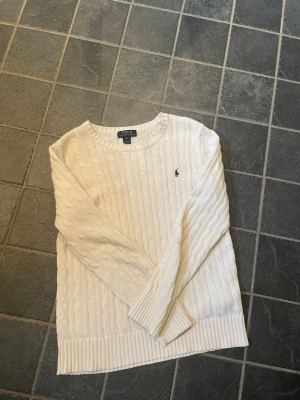 Vit kabelstickad tröja Polo Ralph Lauren - Kabelstickad vit tröja från Polo Ralph Lauren med rund halsringning och diskret broderad logga på bröstet. Tröjan har långa ärmar och breda ribbade muddar vid ärmslut och nederkant. Perfekt för en klassisk och stilren look.