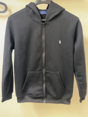 Svart hoodie från Polo Ralph Lauren - Klassisk svart hoodie med dragkedja från Polo Ralph Lauren. Har huva, två fickor framtill och den ikoniska vita logon broderad på bröstet. Tillverkad i mjukt bomullsmaterial som är skönt att bära. Perfekt för en avslappnad och stilren look.