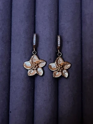 Guldiga blomörhängen med stenar - Säljer ett par snygga örhängen i guldfärgad metall med blomformade hängen. Örhängena har små gnistrande stenar på bygeln och detaljerade blomblad. Perfekta för dig som vill ha något unikt och trendigt i smyckeskrinet.