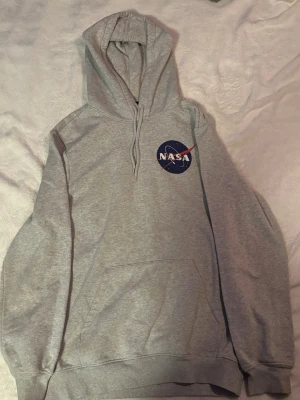 Grå NASA hoodie - Grå hoodie med NASA-logga tryckt på bröstet och en stor NASA-logga på ryggen. Hoodien har huva med en klassisk magficka.