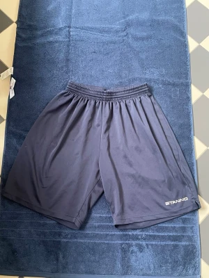 Mörkblå shorts från Stanno - Säljer ett par mörkblå shorts från Stanno med elastisk midja och enkel design. Tillverkade i ett lätt och svalt syntetmaterial, perfekta för sport eller träning. Diskret logga på ena benet och ledig passform.