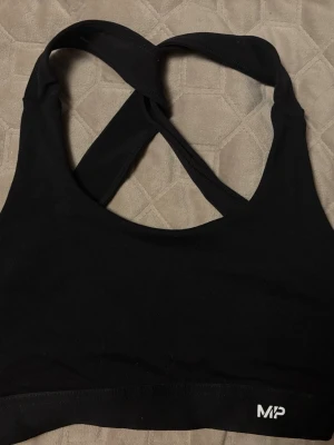 Svart sport-bh från MP - Säljer en svart sport-bh från MP med korsade axelband i ryggen. Bh:n har en enkel och stilren design utan vaddering och är tillverkad i ett stretchigt material som passar perfekt för träning. Diskret MP-logga framtill.