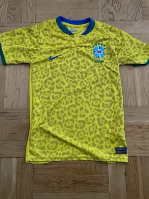 Brasilien Nike matchtröja gul leopard - Säljer en Brasilien matchtröja från Nike i starkt gul färg med leopardmönster på framsidan och ärmarna. Tröjan har blå krage och gröna muddar, samt Brasilien-emblem och Nike-logga på bröstet. Materialet är lätt och andas, perfekt för sport och träning.