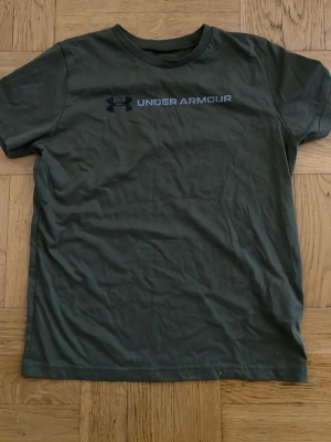 Grön Under Armour t-shirt  - Mörkgrön t-shirt från Under Armour med klassisk logga och text på bröstet. Modellen har korta ärmar och rund halsringning. Tillverkad i mjuk bomull och har en loose passform som ger en avslappnad look.