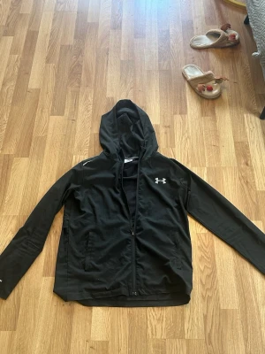Svart träningsjacka från Under Armour - Svart träningsjacka med huva från Under Armour. Jackan har dragkedja framtill, två fickor och reflekterande detaljer vid axlarna. Tillverkad i lätt syntetmaterial, perfekt för utomhusaktiviteter.