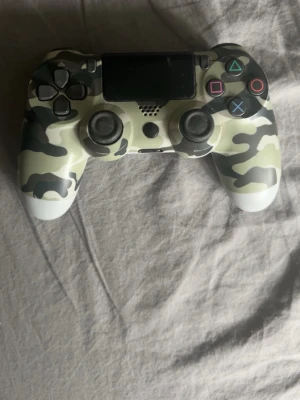 Camo DualShock 4 handkontroll PS4 - Snygg PlayStation 4 DualShock 4 handkontroll från Sony med grått och vitt kamouflagemönster på framsidan och vit baksida. Kontrollern har klassiska PlayStation-knappar med färgade symboler och greppvänlig form i plast. Perfekt för gaming på PS4.