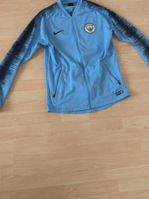 Nike Manchester City träningströja  - Ljusblå träningsjacka från Nike med Manchester Citys klubbmärke på bröstet. Jackan har hel dragkedja, två fickor med dragkedja och svarta detaljer längs ärmarna. Tillverkad i Dri-FIT-material som andas och passar perfekt till träning.