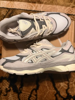 ASICS gråvita sneakers mesh & mocka - Snygga sneakers från ASICS i grått, vitt och beige. Ovandelen är i mesh och mocka med klassiska ASICS-stripes på sidan. Skorna har en chunky sula i vitt och grått och låga snörningar. Perfekta för dig som gillar retrovibbar och streetstyle.