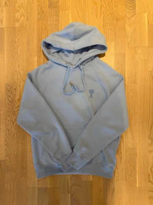 Ljusblå hoodie från Ami Paris - Snygg ljusblå hoodie från Ami Paris med det ikoniska hjärta-A broderiet på bröstet. Hoodien har dragsko i huvan, ribbade muddar och en klassisk känguruficka. Tillverkad i mjuk bomull för maximal komfort.