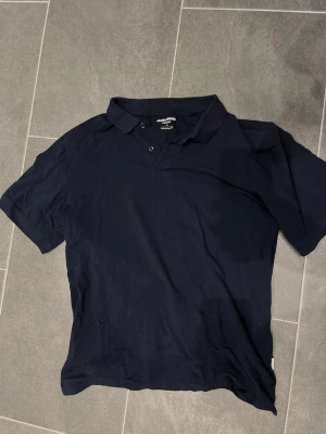 Mörkblå pikétröja från Jack & Jones - Klassisk mörkblå pikétröja från Jack & Jones med krage och två knappar framtill. Tillverkad i mjuk bomull som känns skön mot huden. Perfekt till jeans eller shorts för en enkel och stilren look.