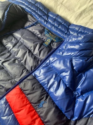 Blå dunjacka från Polo Ralph Lauren - Snygg blå dunjacka från Polo Ralph Lauren med quiltad design och dragkedja framtill. Jackan har en röd och marinblå rand över bröstet samt broderad logga på vänstersidan. Perfekt för kalla vinterdagar och riktigt stilren look.