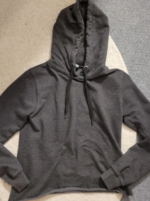 Mörkgrå hoodie med huva - Mysig mörkgrå hoodie med stor huva och svarta dragsnören. Tröjan har långa ärmar och ribbade muddar vid ärmslut. Perfekt för chill dagar och enkel att matcha med jeans eller mjukisbyxor. 