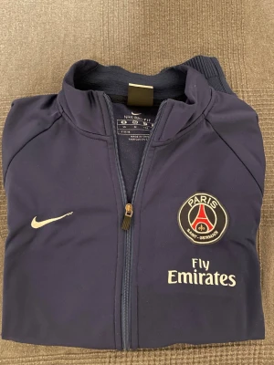 Mörkblå PSG träningsjacka Nike - Snygg mörkblå träningsjacka från Nike med Paris Saint-Germain klubbmärke och Fly Emirates-tryck på bröstet. Jackan har hel dragkedja, ståkrage och två sidofickor. Tillverkad i ett lätt och funktionellt material, perfekt för träning eller sport.