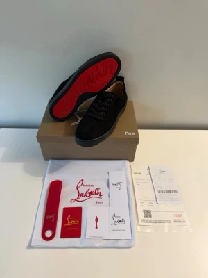 Christian Louboutin svarta sneakers - Snygga svarta sneakers från Christian Louboutin med klassisk röd sula och lågt skaft. Skorna har ovandel i svart textil och läderdetaljer, samt svarta skosnören. Ikonisk design med märkets logga på sulan. Perfekta för dig som gillar exklusiva och trendiga sneakers.