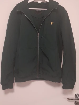 Grön softshell jacka från Lyle & Scott - Grön softshelljacka från Lyle & Scott med huva och hel dragkedja. Jackan har en gul broderad logga på bröstet, tre stora fickor framtill och ribbade muddar vid ärmsluten. Tillverkad i softshell material som passar perfekt för blåsiga dagar.
