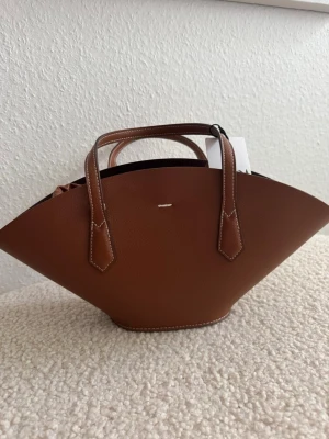Brun handväska  med unik form - Snygg brun handväska med en unik, bred och bågformad design. Väskan har dubbla handtag, vita kontrastsömmar och en innerpåse med dragkedja och karbinhake. Perfekt för dig som vill sticka ut med en trendig accessoar.