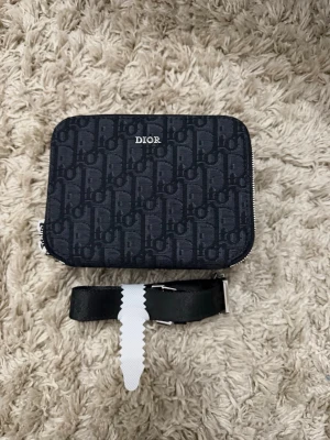 Svart Dior handväska med monogram - Snygg svart handväska från Dior med klassiskt monogrammönster och silverfärgad dragkedja. Väskan har en avtagbar axelrem i svart läder och en kompakt, rektangulär form. Perfekt för dig som gillar exklusiva accessoarer med tydlig logga.