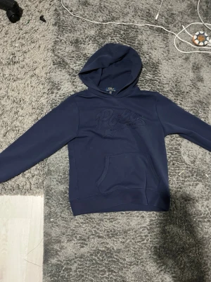 Mörkblå hoodie från Polo Ralph Lauren - Snygg mörkblå hoodie från Polo Ralph Lauren med broderad logga på bröstet. Klassisk känguruficka framtill och stor huva. Tillverkad i mjukt bomullsmaterial som känns skönt mot huden. Perfekt för en avslappnad och trendig look.