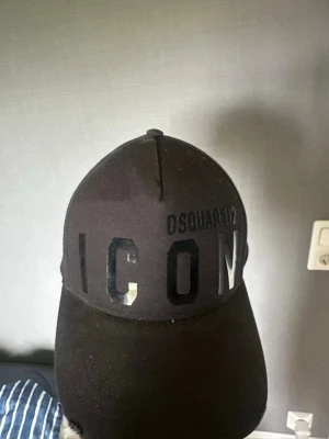 Svart ICON keps från Dsquared2 - Säljer en svart keps från Dsquared2 med stor text 'ICON' framtill och broderad logga. Klassisk böjd skärm och justerbar rem bak. Tillverkad i bomull för en bekväm och snygg look.