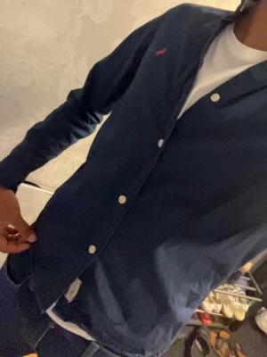 Mörkblå skjorta med knappar - Snygg mörkblå skjorta med vita knappar och diskret röd logga på bröstet. Skjortan har lång ärm och klassisk krage, perfekt till jeans eller chinos. Tillverkad i bomull för en skön känsla och stilren look.