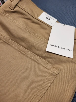 Beige chinos från These Glory Days 54 L - Snygga beige chinos från These Glory Days i storlek 54. Tillverkade i ekologisk bomull med klassisk passform, framfickor och bälteshällor. Perfekta för en clean och stilren look.